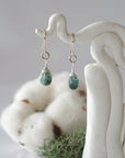 Detailed front view of handcrafted emerald sterling silver drop earrings with natural faceted emerald, artisan wire wrapping, hanging from a display – Designs by Nature Gems