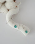 Turquoise sterling silver drop earrings with faceted natural turquoise stones and wire wrapping, laying flat on a display – Designs by Nature Gems