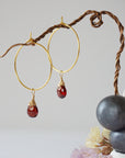 Close-up of handcrafted garnet hoop drop earrings in gold plated brass, featuring wire wrapped natural red garnet gemstones on a jewelry display