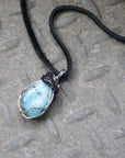 Men's aquamarine necklace laying flat, artisan wire wrapped pendant in black and white gold finish, black leather cord