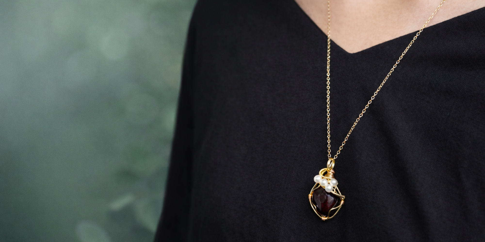 Person wearing a pearl and garnet gold necklace with a pendant against a blurred green background