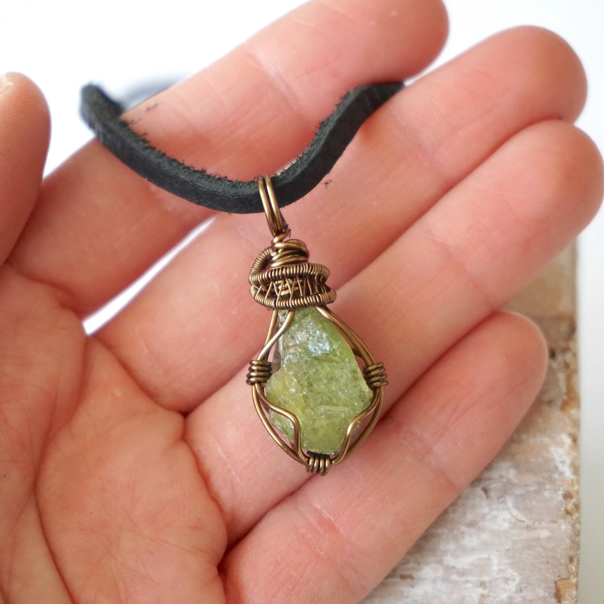 Peridot pendant for men in hand for size reference – natural green crystal, bronze copper wire, black leather cord, August birthstone.