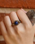 Hand wearing a gold ring with a blue stone against a brick wall background