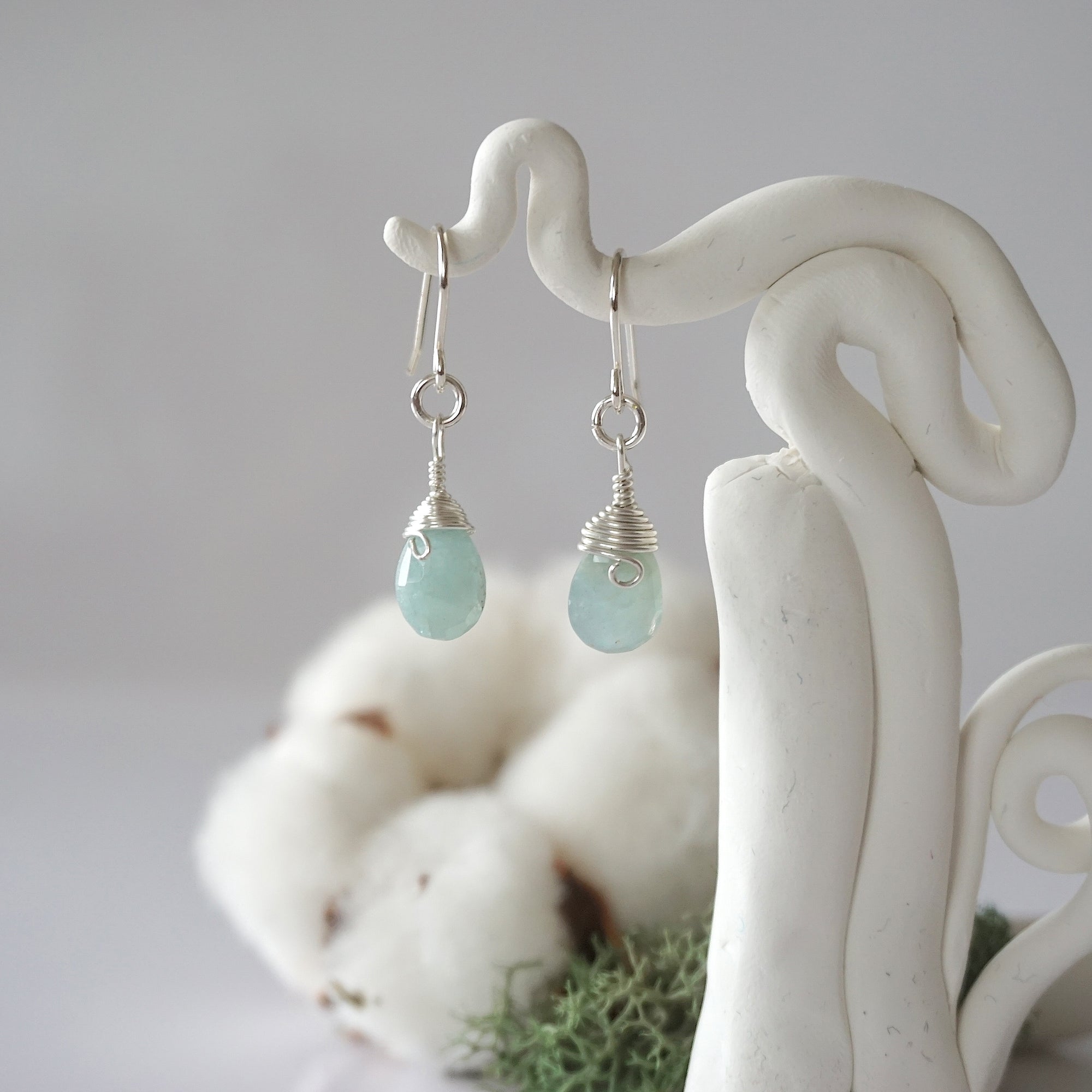 Detailed front view of handcrafted aquamarine sterling silver drop earrings with natural faceted aquamarine, artisan wire wrapping, hanging from a display – Designs by Nature Gems