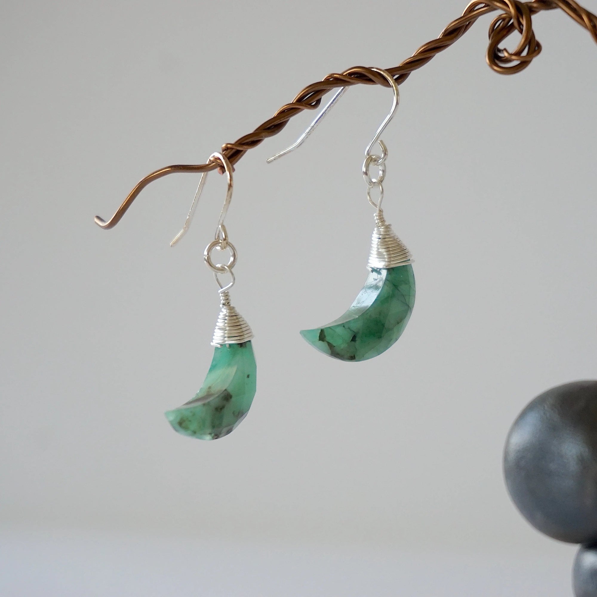 Side angle view of emerald moon drop earrings showing sterling silver artisan wire wrapping and faceted emerald detail.