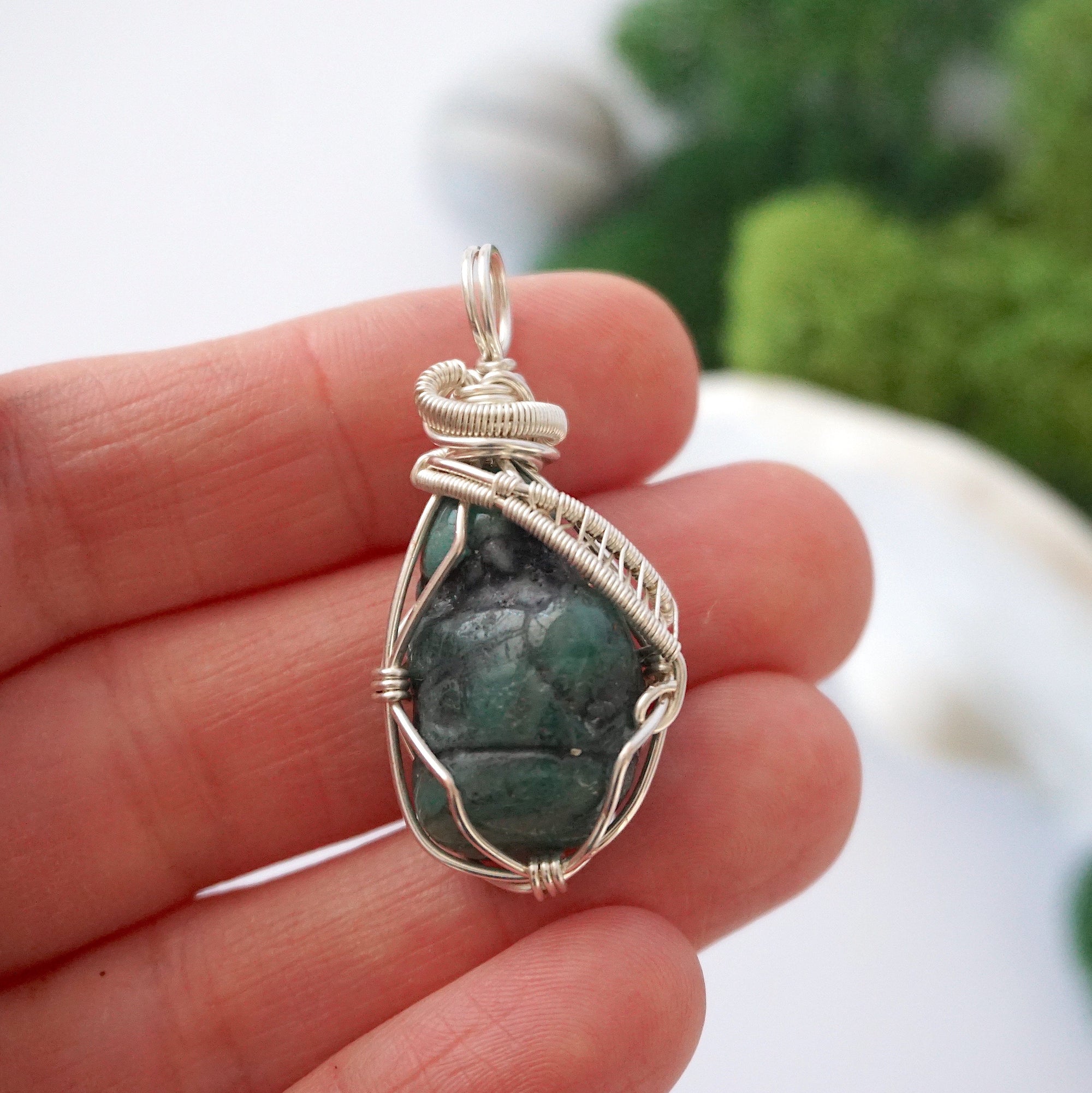 Natural raw emerald pendant shown in hand for size reference, wrapped in detailed soft silver copper wire.