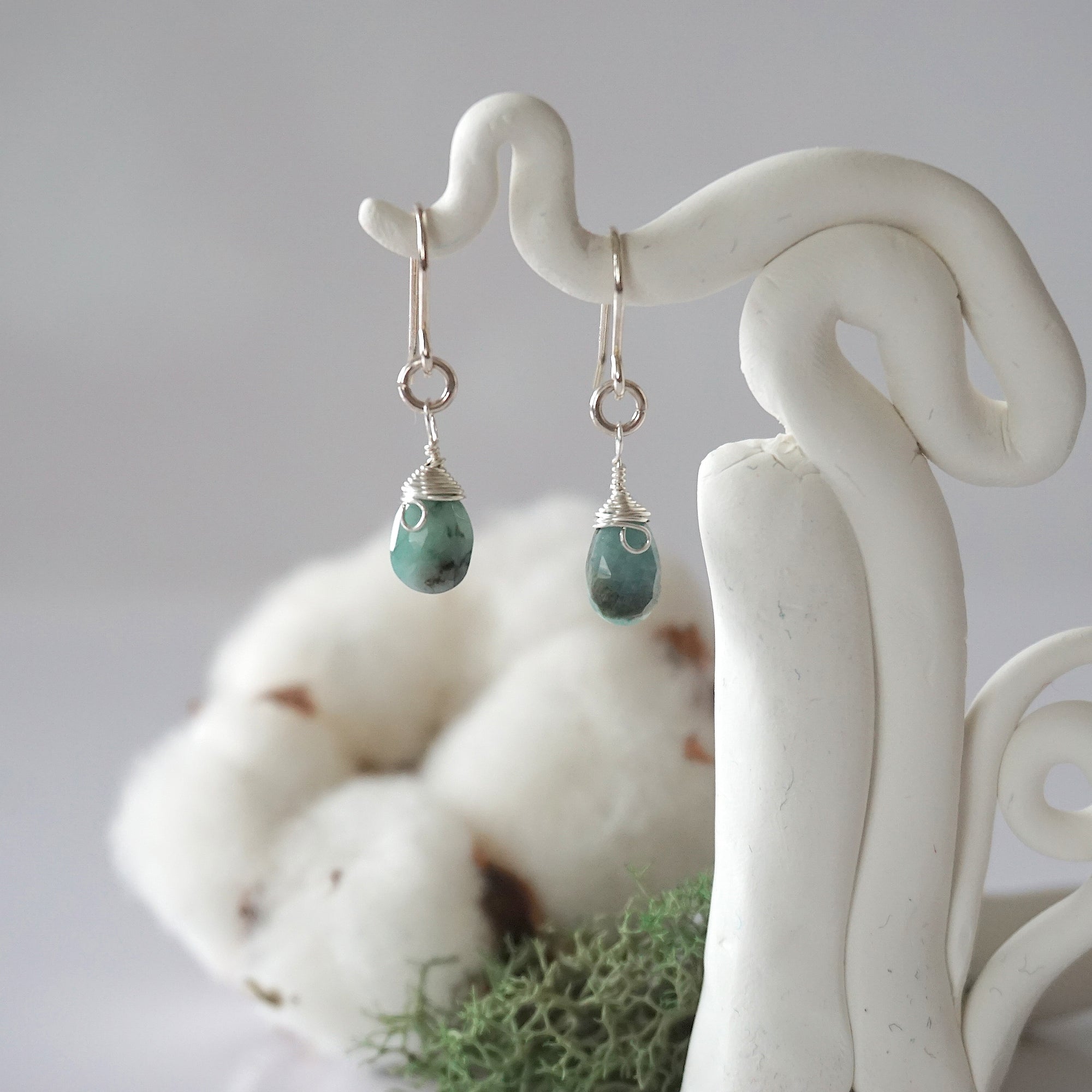 Detailed front view of handcrafted emerald sterling silver drop earrings with natural faceted emerald, artisan wire wrapping, hanging from a display – Designs by Nature Gems