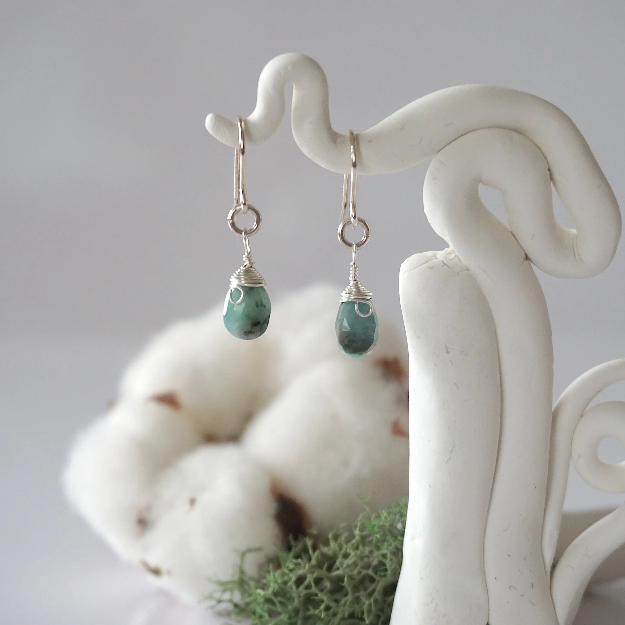 Detailed front view of handcrafted emerald sterling silver drop earrings with natural faceted emerald, artisan wire wrapping, hanging from a display – Designs by Nature Gems