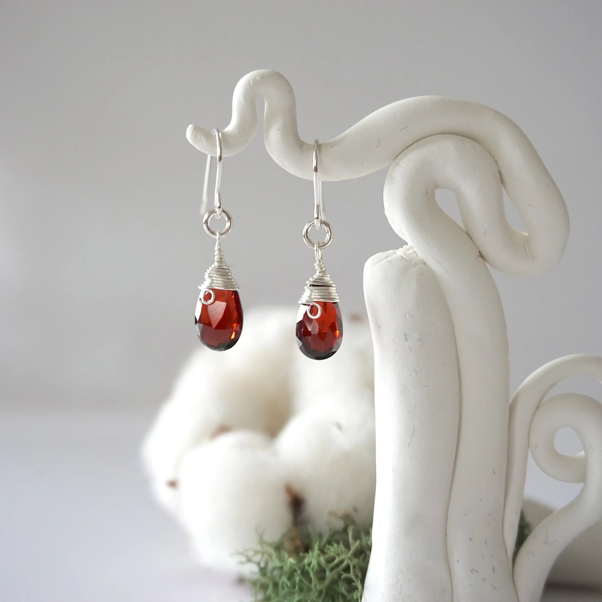 Detailed front view of handcrafted garnet sterling silver drop earrings with natural faceted garnet, artisan wire wrapping, hanging from a display – Designs by Nature Gems