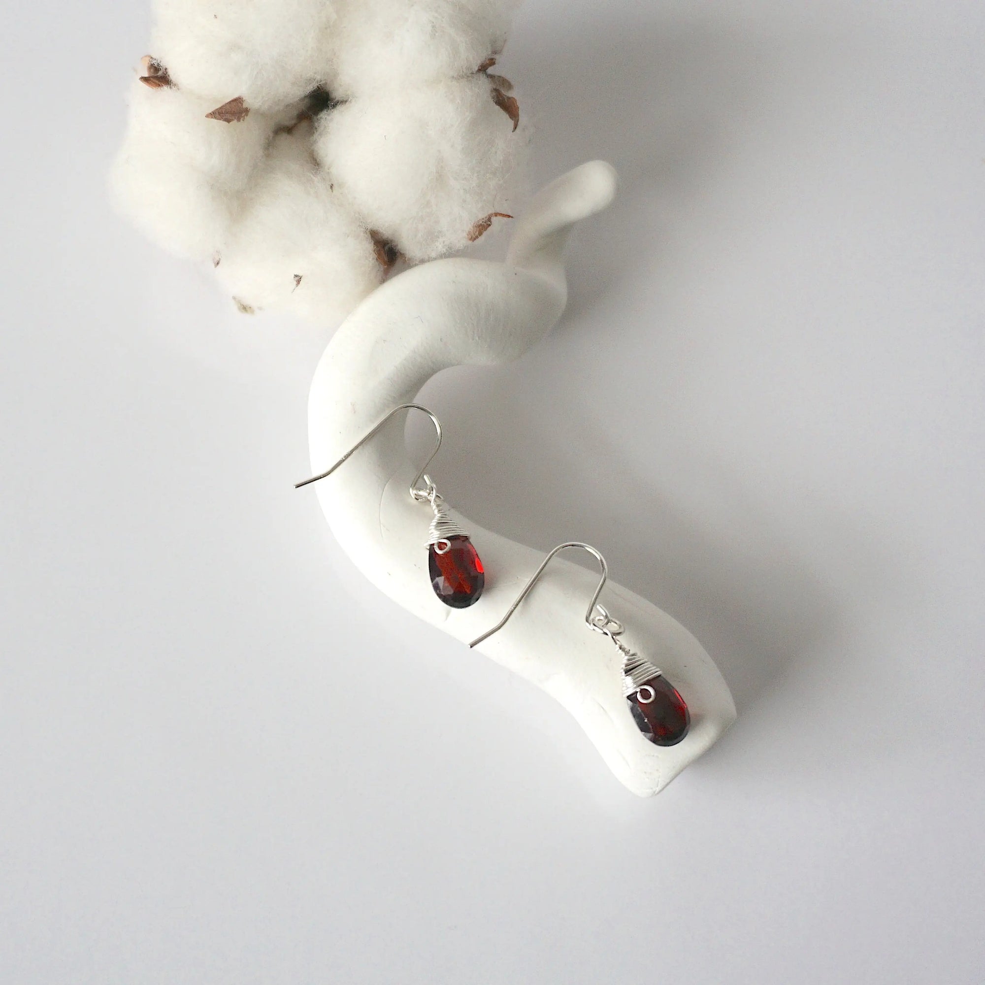 Garnet sterling silver drop earrings with faceted natural garnet stones and wire wrapping, laying flat on a display – Designs by Nature Gems
