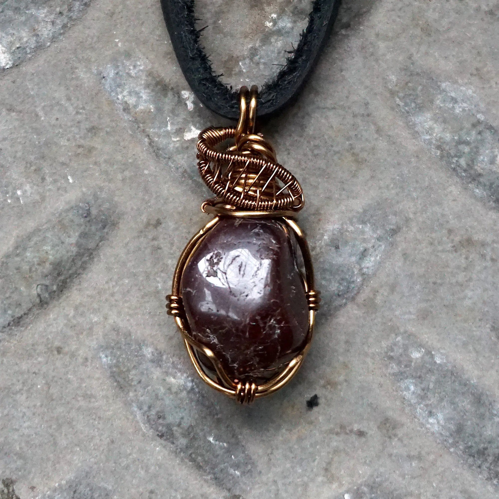 Men’s garnet wire wrapped necklace in antique bronze copper on a black leather cord, shown laying flat to highlight the natural garnet pendant