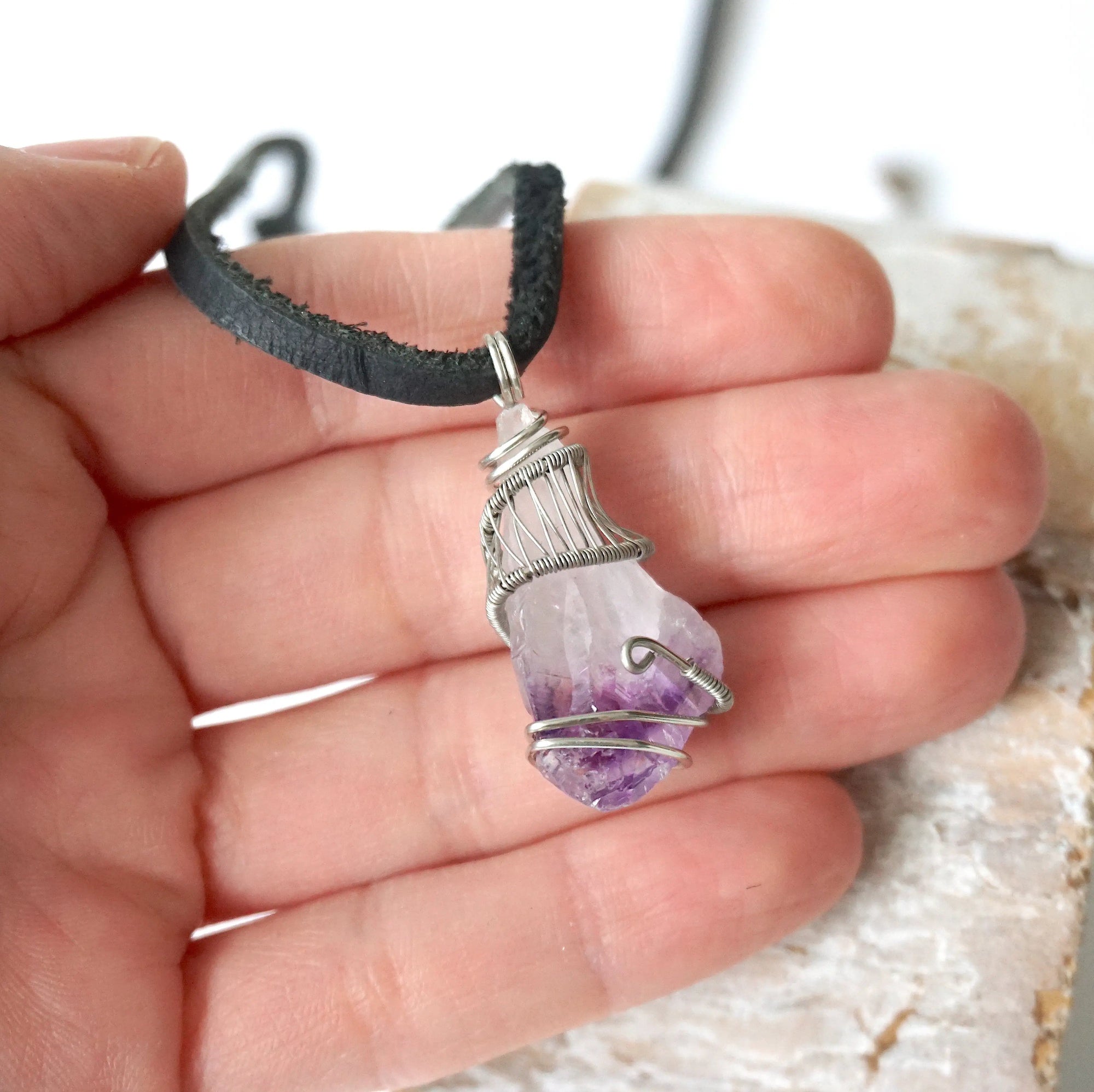 Men’s raw amethyst pendant necklace on black leather cord shown in hand for size reference, white gold plated copper wire wrap