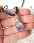 Men’s raw amethyst pendant necklace on black leather cord shown in hand for size reference, white gold plated copper wire wrap
