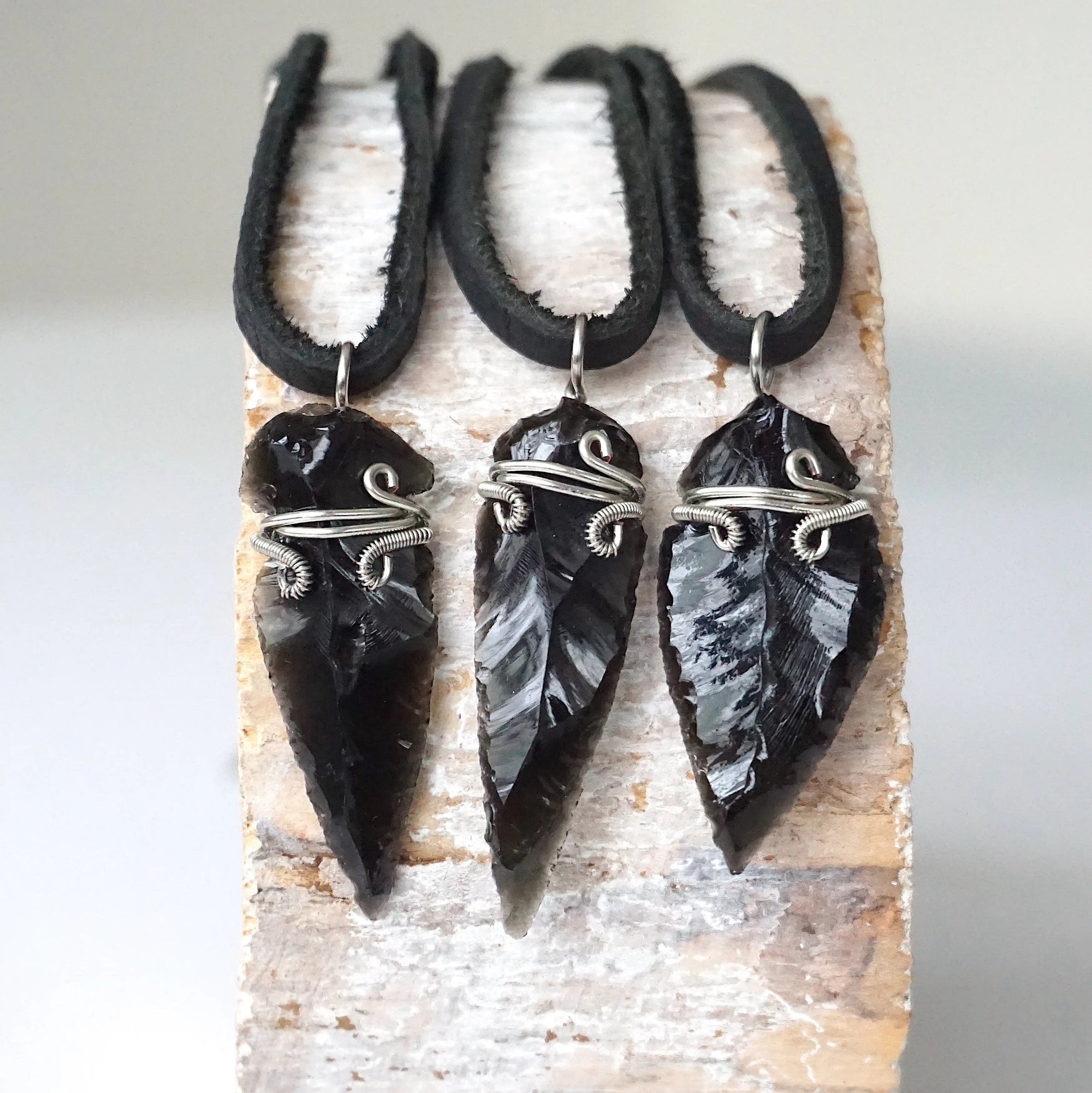 Three men’s black obsidian arrowhead pendants on black leather cords, showing natural crystal variations, artisan jewelry by Designs by Nature Gems.