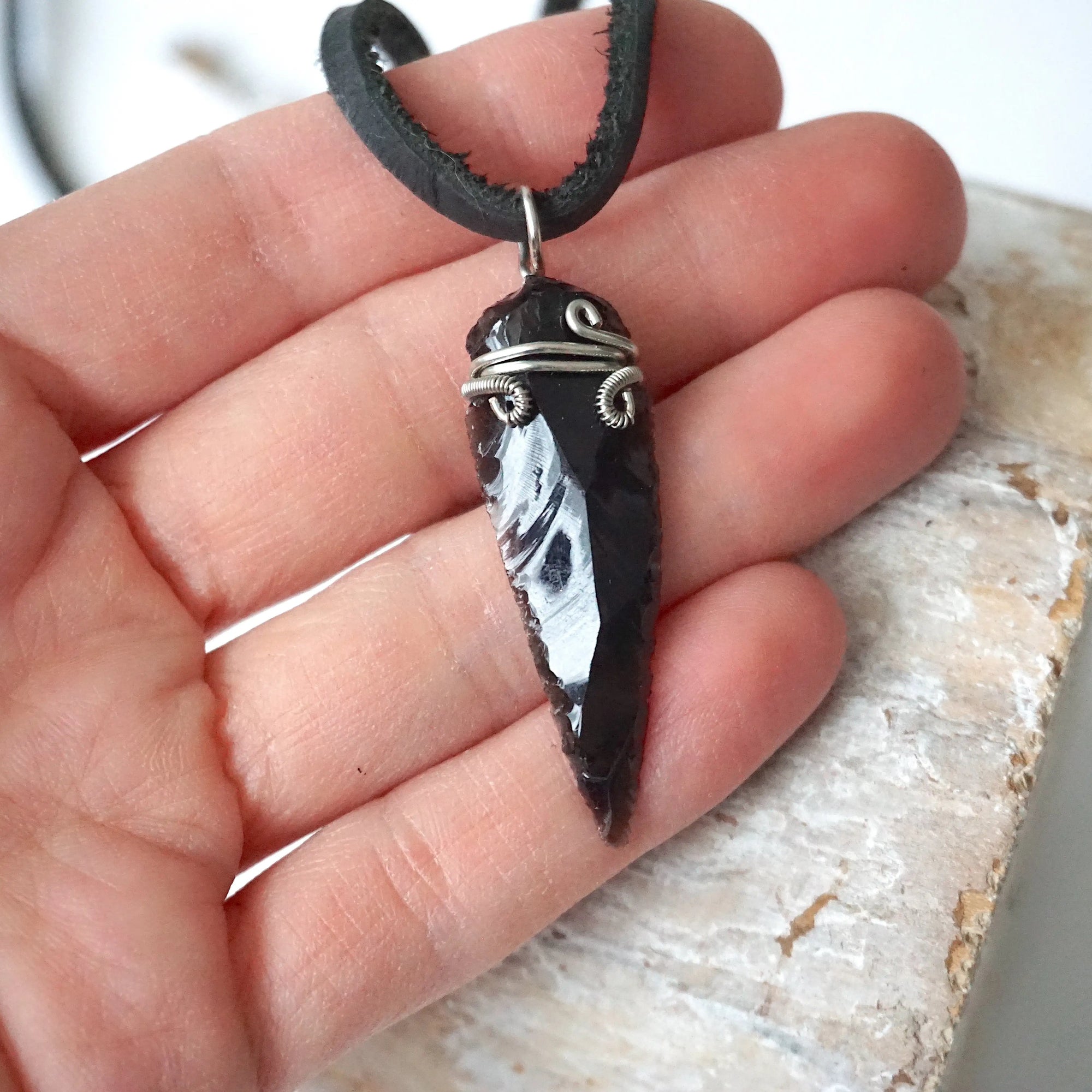 Black obsidian arrowhead necklace for men shown on black leather cord in a hand for size reference, natural crystal jewelry by Designs by Nature Gems.