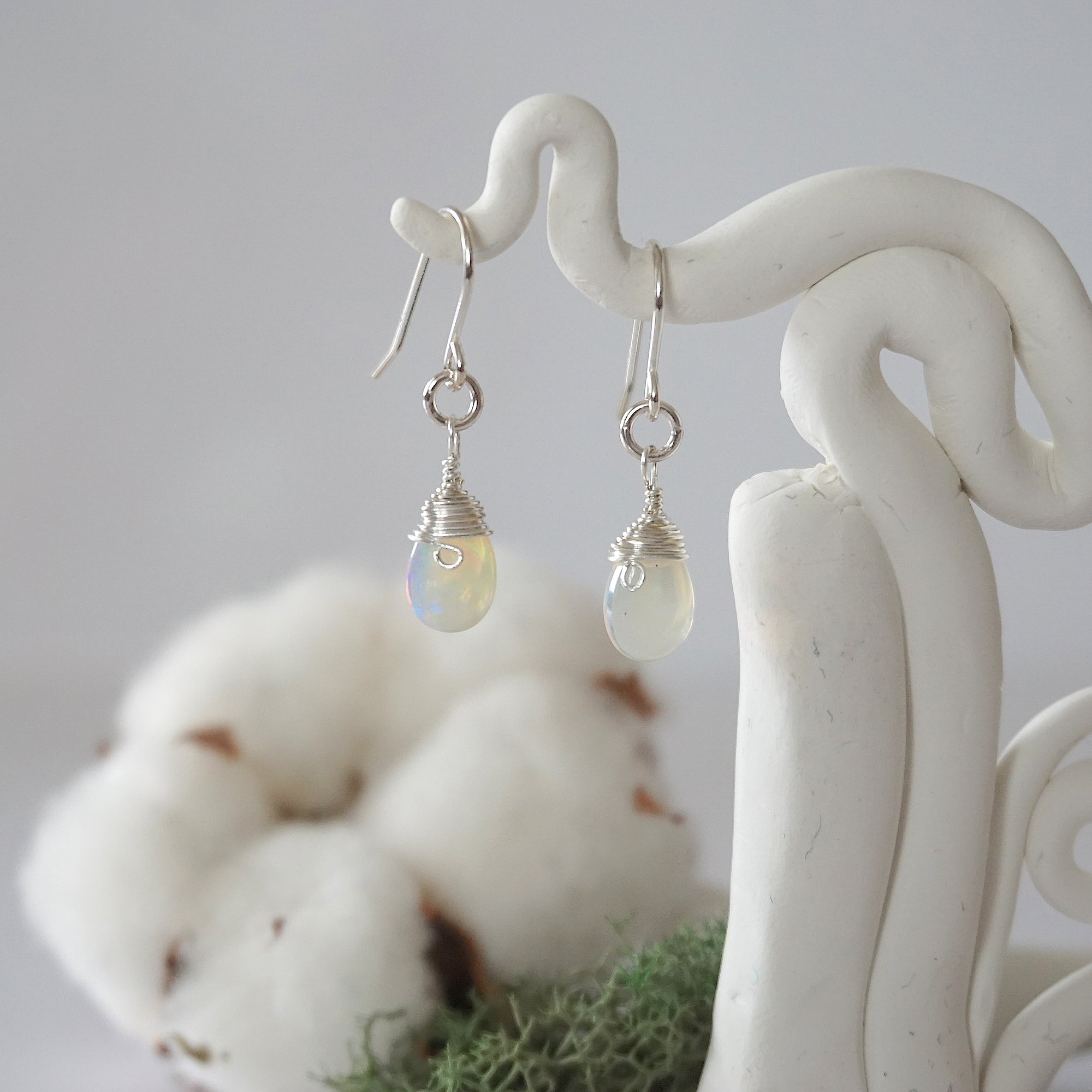 Detailed front view of handcrafted opal sterling silver drop earrings with natural faceted opal, artisan wire wrapping, hanging from a display – Designs by Nature Gems