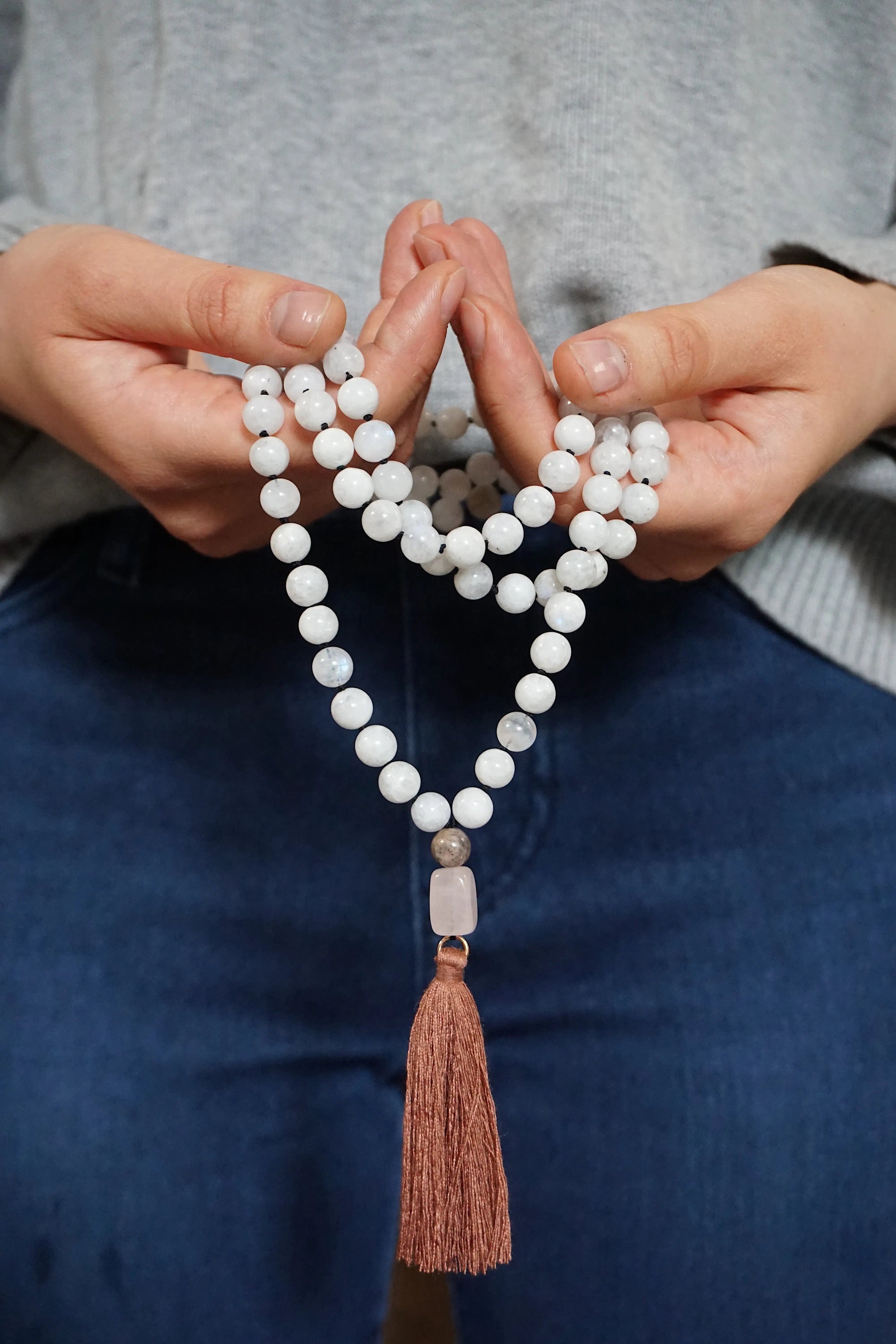 Rainbow moonstone mala necklace with 108 beads and rose quartz guru bead held in hands, knotted silk cord meditation necklace with brown tassel.