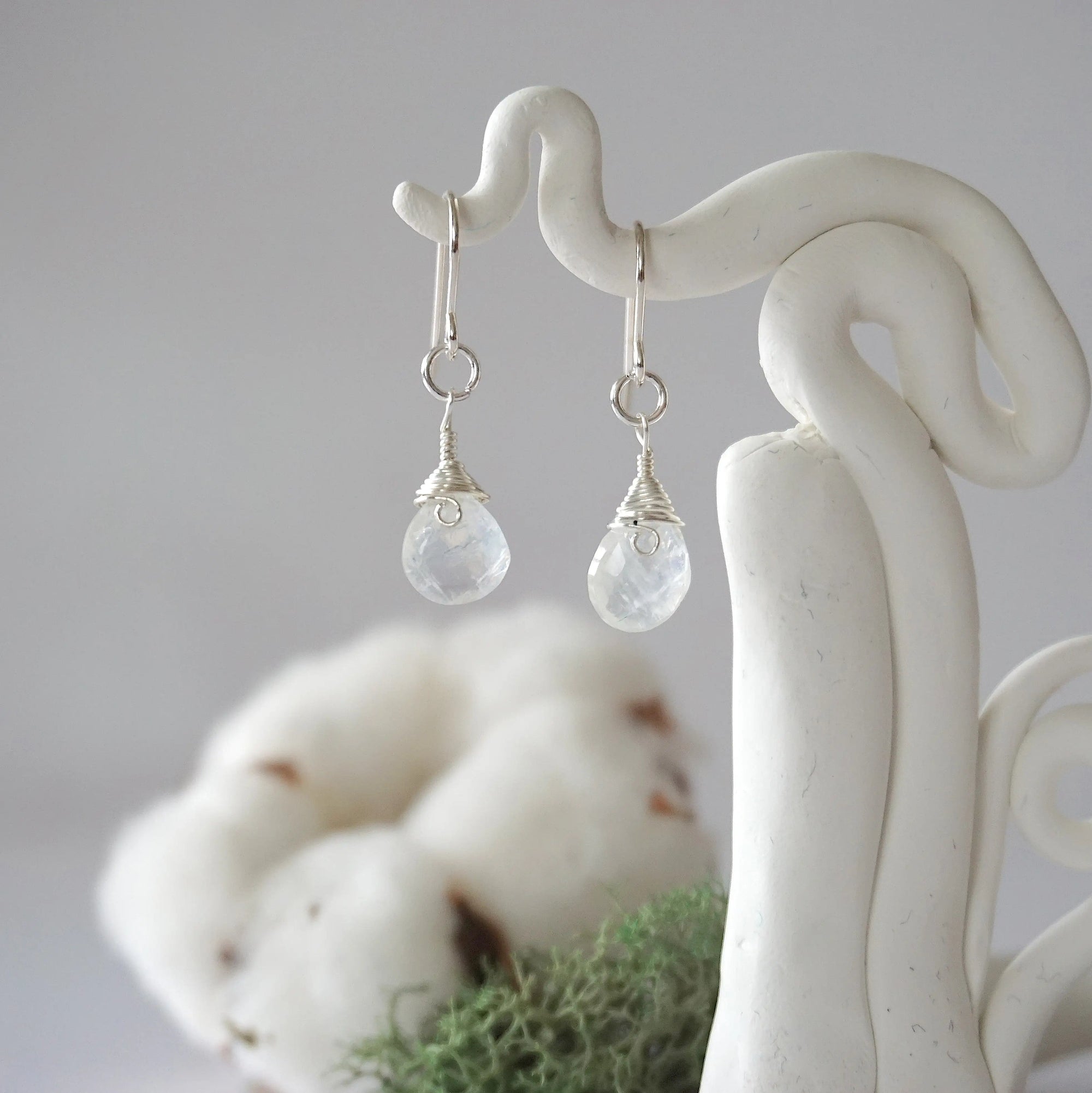 Detailed front view of handcrafted rainbow moonstone sterling silver drop earrings with natural faceted rainbow moonstone, artisan wire wrapping, hanging from a display – Designs by Nature Gems