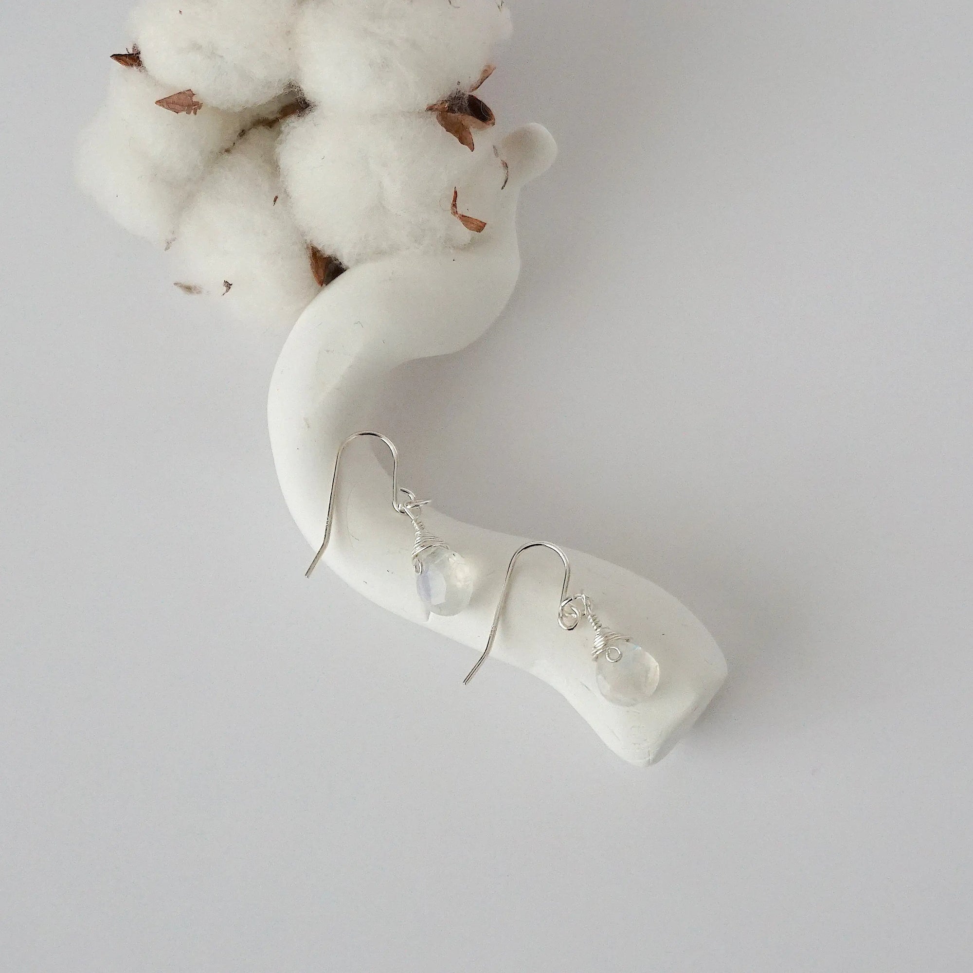 Rainbow moonstone sterling silver drop earrings with faceted natural rainbow moonstone stones and wire wrapping, laying flat on a display – Designs by Nature Gems