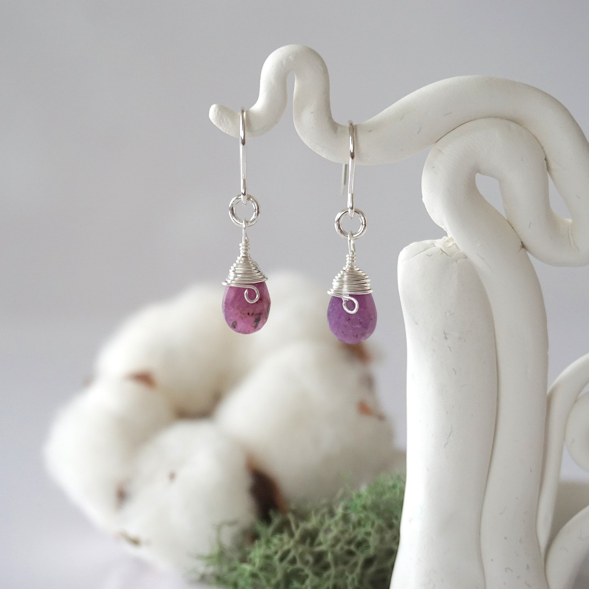 Detailed front view of handcrafted ruby sterling silver drop earrings with natural faceted ruby, artisan wire wrapping, hanging from a display – Designs by Nature Gems