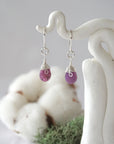 Detailed front view of handcrafted ruby sterling silver drop earrings with natural faceted ruby, artisan wire wrapping, hanging from a display – Designs by Nature Gems