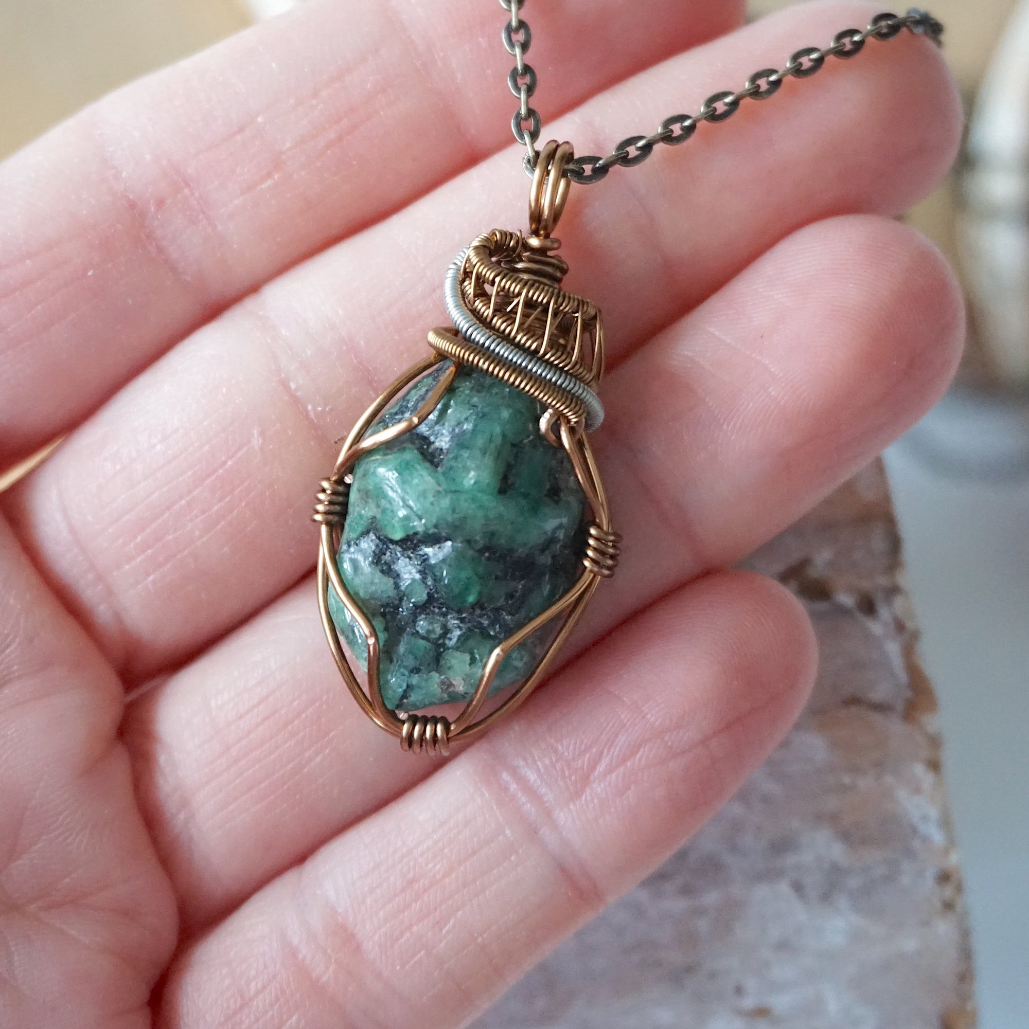 Raw emerald pendant shown in hand for size reference – natural green crystal, bronze and silver copper wire, unisex birthstone jewelry by Designs by Nature Gems.