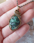 Raw emerald pendant shown in hand for size reference – natural green crystal, bronze and silver copper wire, unisex birthstone jewelry by Designs by Nature Gems.