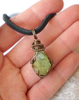 Peridot pendant for men in hand for size reference – natural green crystal, bronze copper wire, black leather cord, August birthstone.