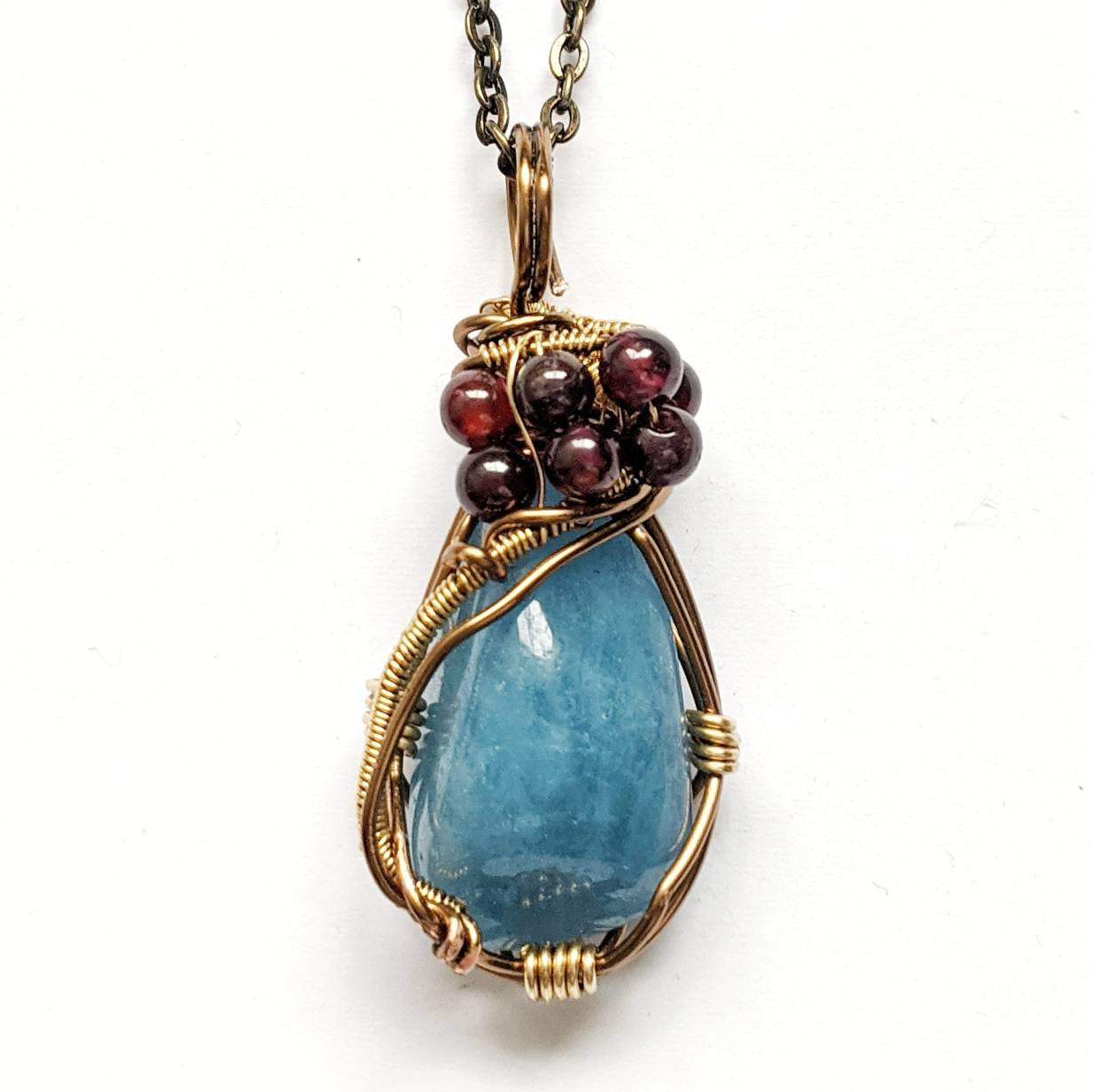 Angled view of a wire wrapped aquamarine pendant necklace with garnet accent beads on an antique bronze chain