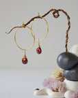 Angled view of gold plated garnet hoop earrings with wire wrapped gemstone drops, showing faceted January birthstone detail
