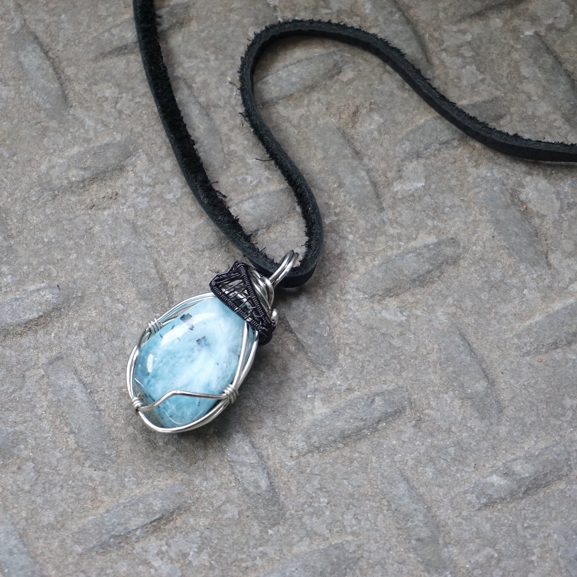 Men's aquamarine necklace laying flat, artisan wire wrapped pendant in black and white gold finish, black leather cord