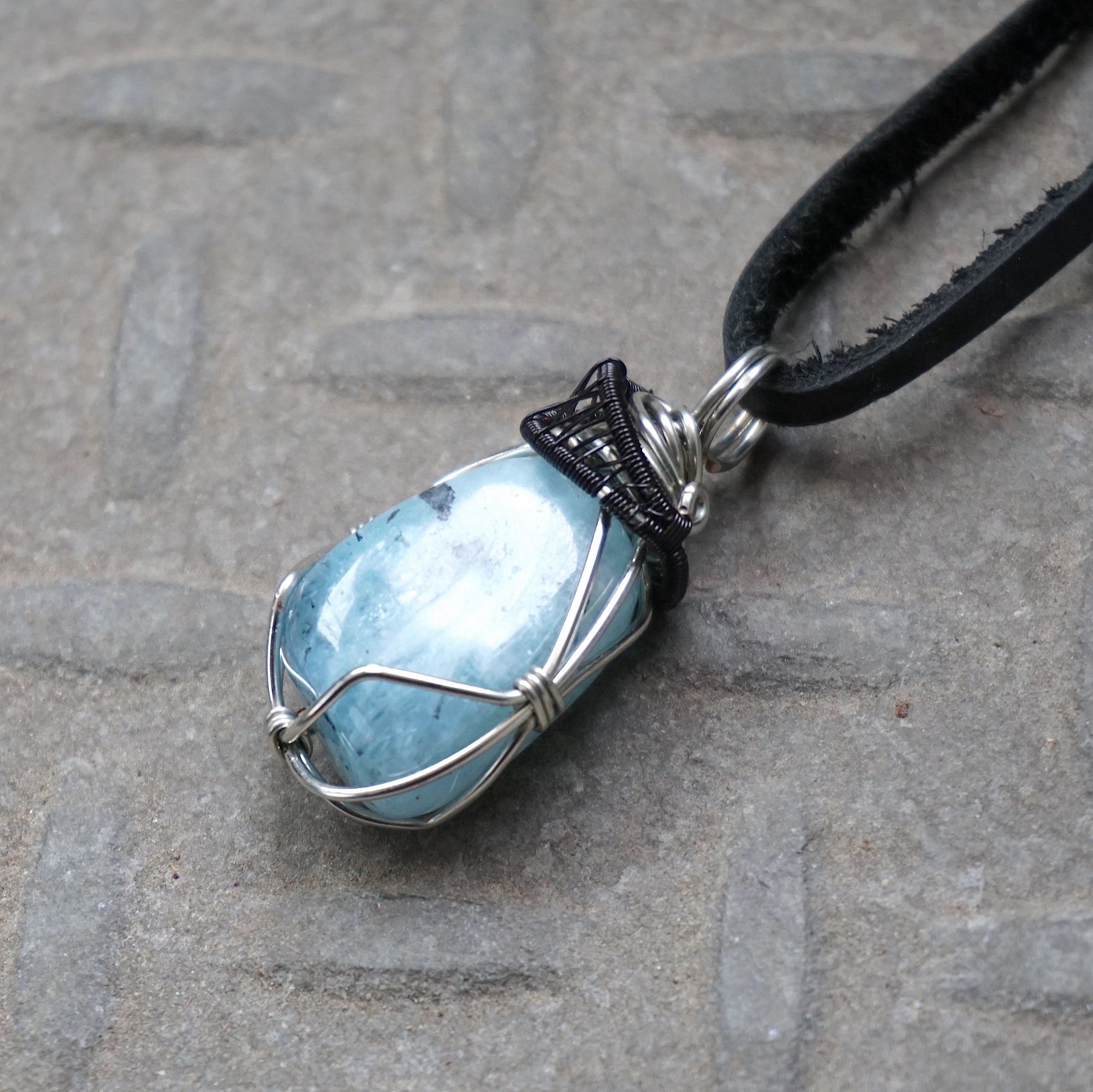Detailed left side angle of men's aquamarine pendant on a black leather cord, black and white gold finish wire wrapping